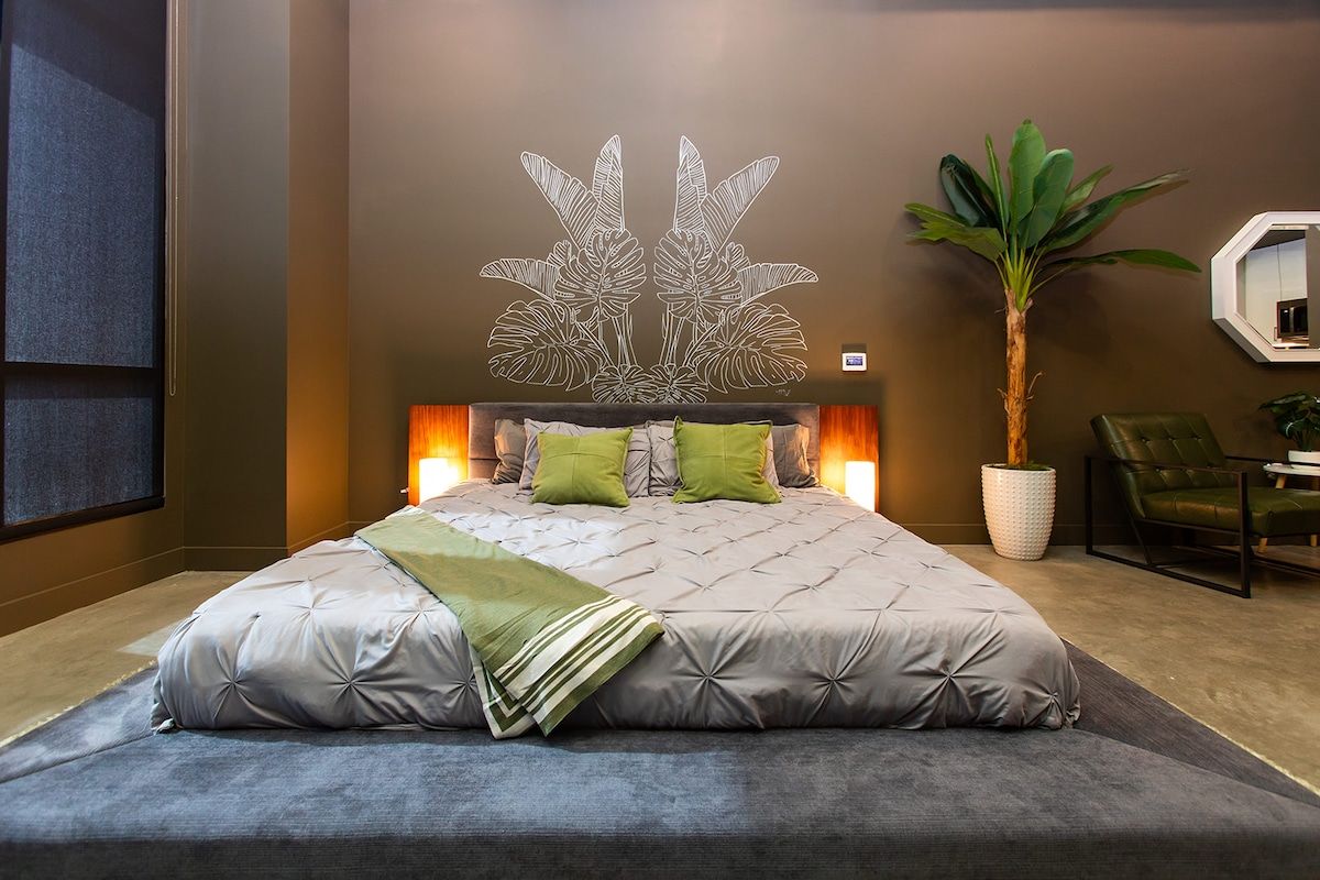 A modern bedroom with a large bed, a wall-mounted mirror, a potted plant, and a green chair.