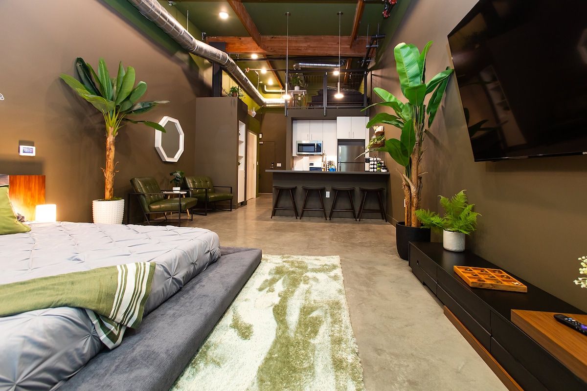 A modern bedroom with a bed, a desk, and a kitchenette in the background.
