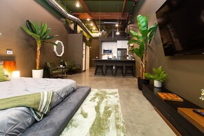A modern bedroom with a bed, a desk, and a kitchenette in the background.