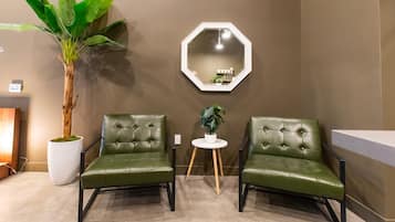 A room with two green leather chairs, a white round table with a plant, a large potted plant, and a hexagonal mirror on the wall.