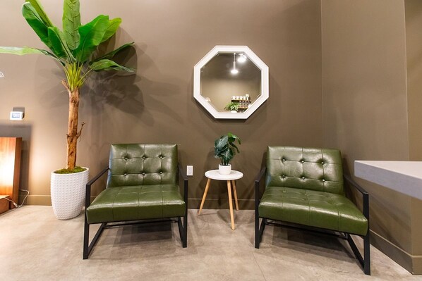 A room with two green leather chairs, a white round table with a plant, a large potted plant, and a hexagonal mirror on the wall.