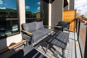Ein Sitzbereich im Freien mit einer Couch und Stühlen, einem kleinen Tisch und Blick auf die Stadt durch große Fenster.