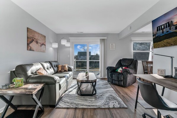 A modern living room with a leather sofa, a black armchair, a wooden coffee table, and a view of the outdoors through large windows.
