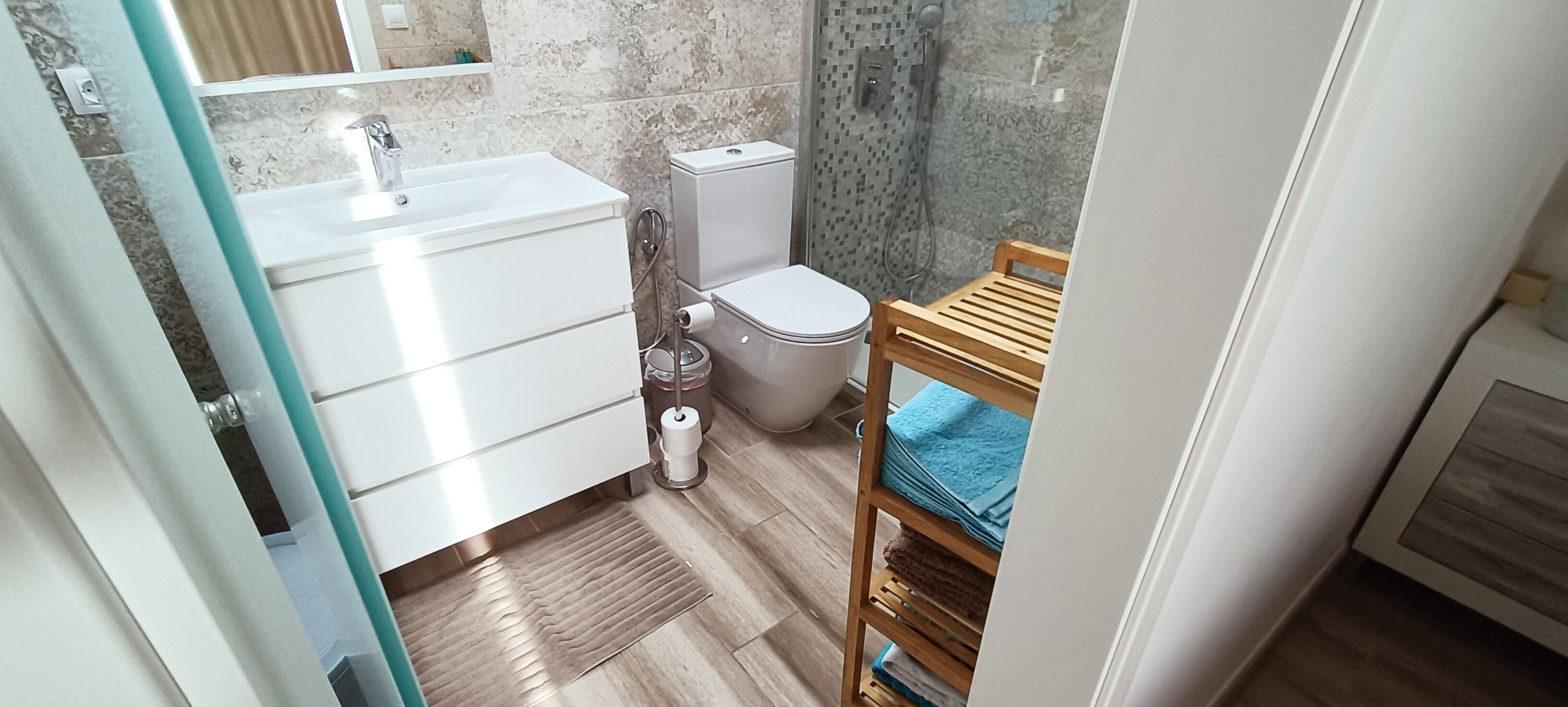 Une salle de bain avec un lavabo, des toilettes et une douche, tous de couleur blanche. On y trouve une chaise en bois et une serviette bleue.