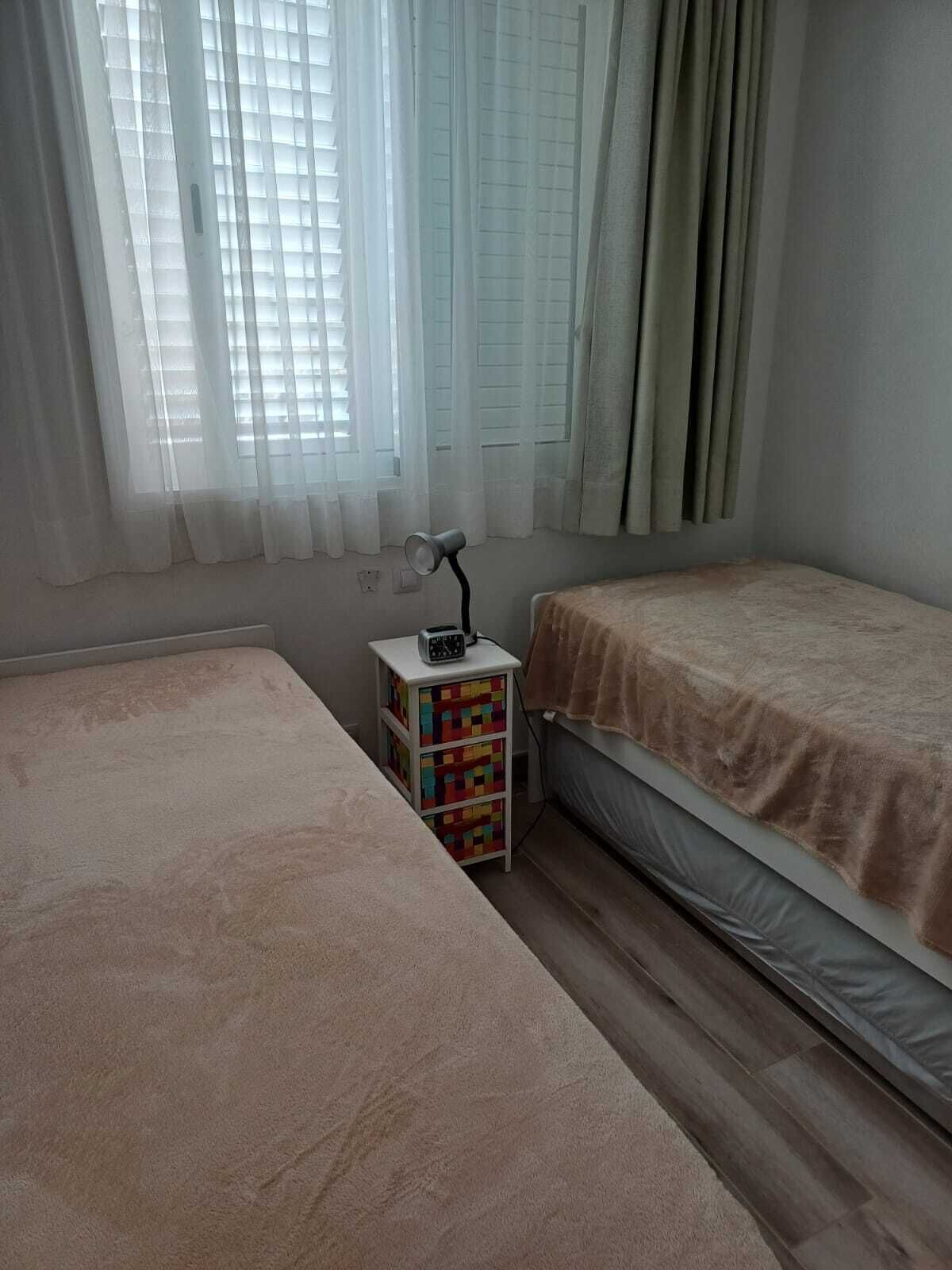 A bedroom with two beds, a bedside table with a lamp, and a window with curtains.