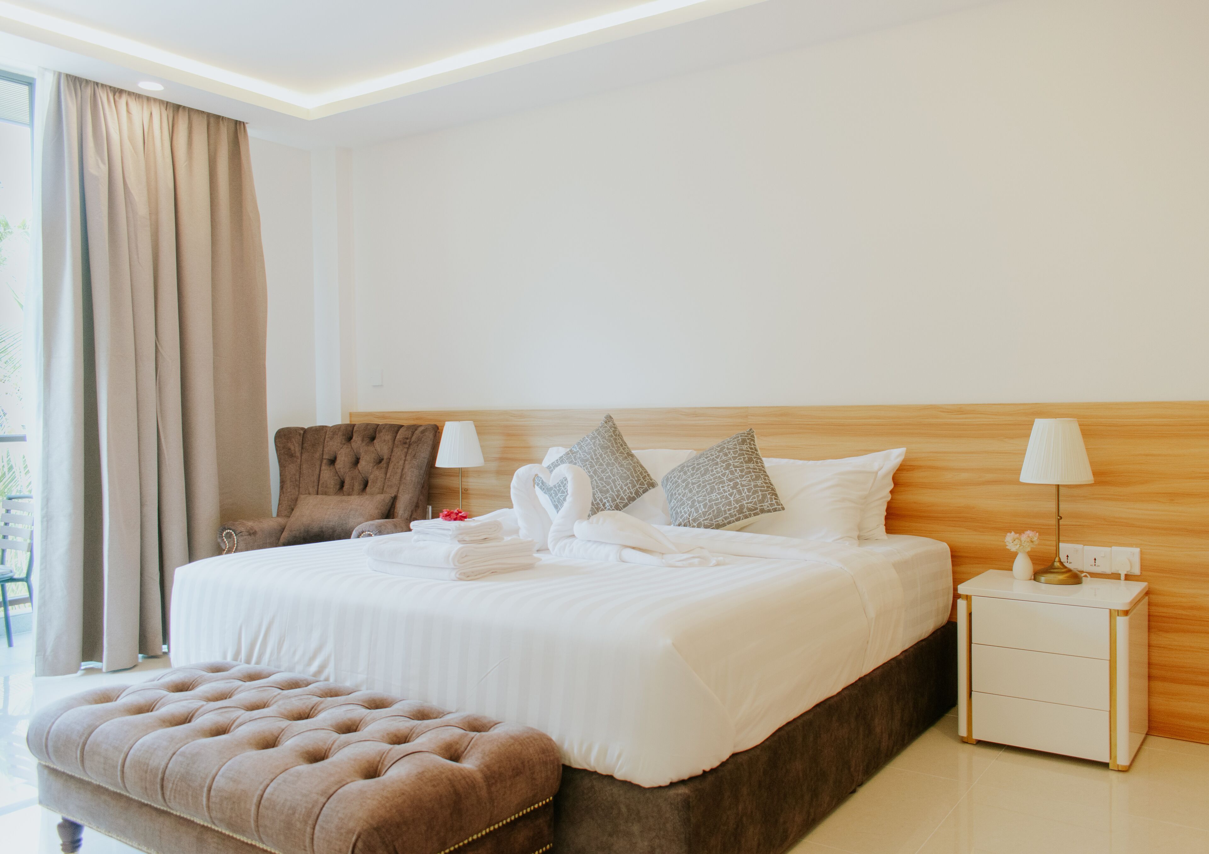 A hotel room with a large bed, a wooden headboard, a brown armchair, a nightstand with a lamp, and a window with curtains.