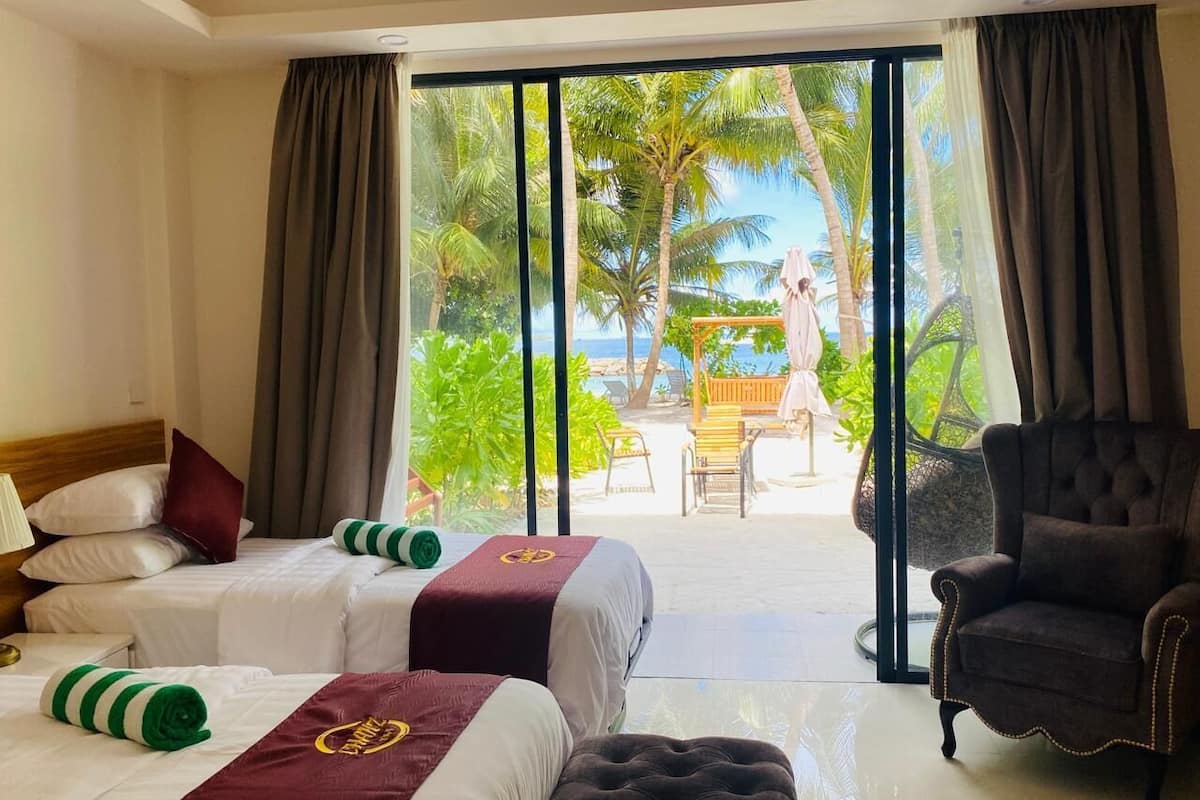 Quarto de hotel com duas camas, uma cadeira e vista para uma praia e palmeiras.