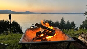 Un foyer en métal avec des bûches et des flammes, entouré d’herbe, avec un lac et des montagnes en arrière-plan.