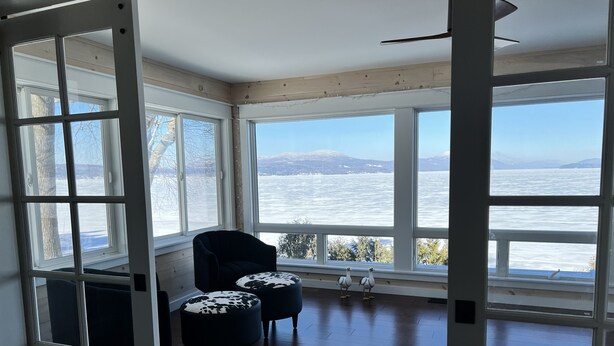 Une pièce dotée d’une grande baie vitrée donnant sur un plan d’eau et des montagnes. On y trouve deux fauteuils noirs et deux poufs à motifs de peau de vache.