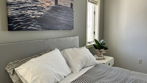 Une chambre à coucher avec un lit, des oreillers, une table de chevet ornée d’une plante, une fenêtre et un tableau représentant un coucher de soleil sur l’eau.