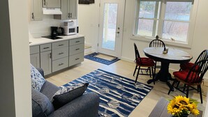 A modern kitchen with white cabinets, a microwave, and a coffee maker. There is a dining area with a table and chairs, a blue rug, and a window with a view of trees.