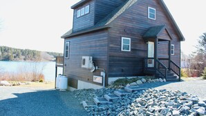 A two-story wooden cabin with a porch and a small staircase, situated by a lake with a gravel driveway and stone steps leading to the entrance.