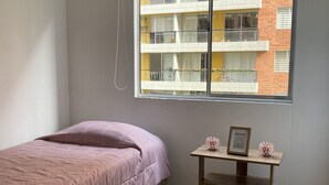 A bedroom with a bed, a small table with a picture frame, a trash bin, and a window with a view of a building. - Hermoso Apartamento Mosquera (guayacan)