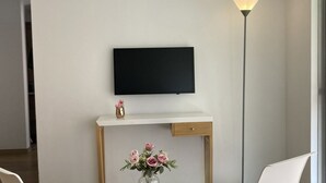 A modern room with a flat-screen TV mounted on the wall, a small wooden desk with a vase of flowers, two white chairs, and a tall floor lamp. - Hermoso Apartamento Mosquera (guayacan)