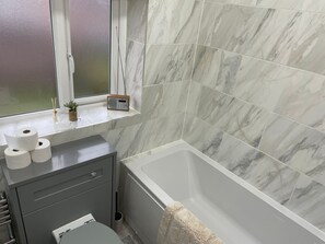 A bathroom with a bathtub, marble tiles, a window with a view, a radio, and a small plant. - Beautiful 3-bed House in Romford (Romford)