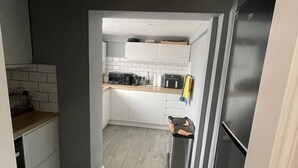 A modern kitchen with grey walls, white cabinets, and a grey floor. There is a black refrigerator on the right and a microwave on the counter. - Beautiful 3-bed House in Romford (Romford)