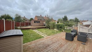 A wooden deck with outdoor furniture, a grassy lawn, and a brick building in the background. - Beautiful 3-bed House in Romford (Romford)