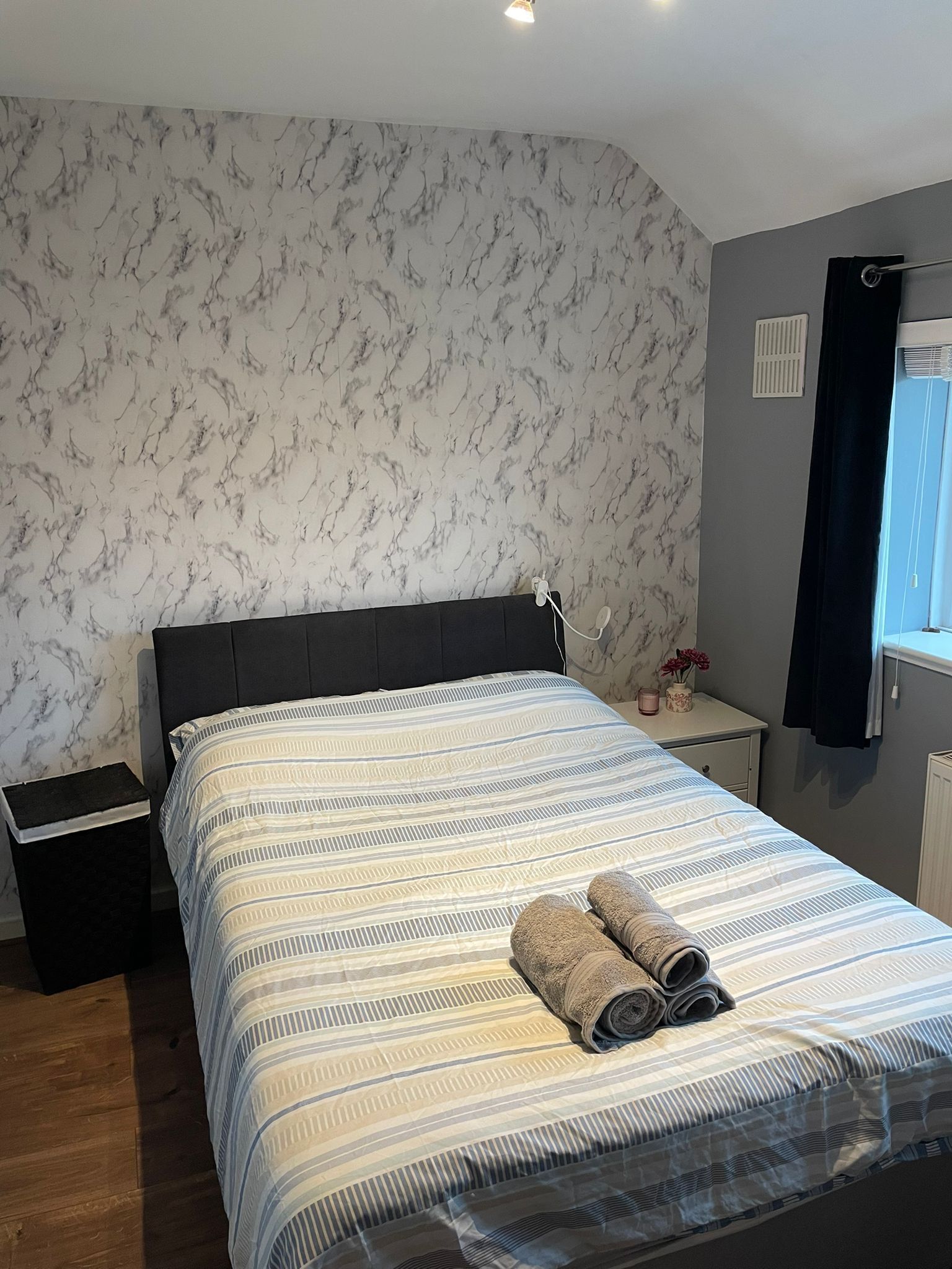 Un dormitorio con cama, mesita de noche, cómoda, ventana con cortinas y pared con diseño de mármol.
