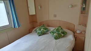 En enkeltseng med to grønne puder med bladmotiv, et natbord med lampe, et spejl og en lille hylde med en glasskål og en rulle køkkenrulle.