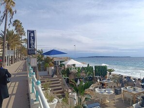 Un restaurante frente al mar con terraza al aire libre, un paseo peatonal y palmeras.