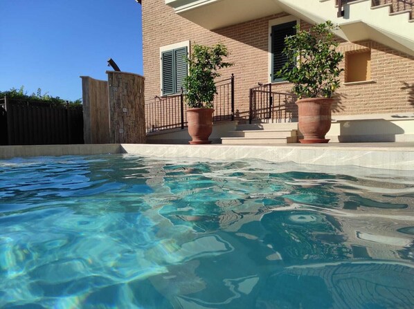 Une piscine à l’eau claire, une maison en briques avec un balcon et deux plantes en pot.