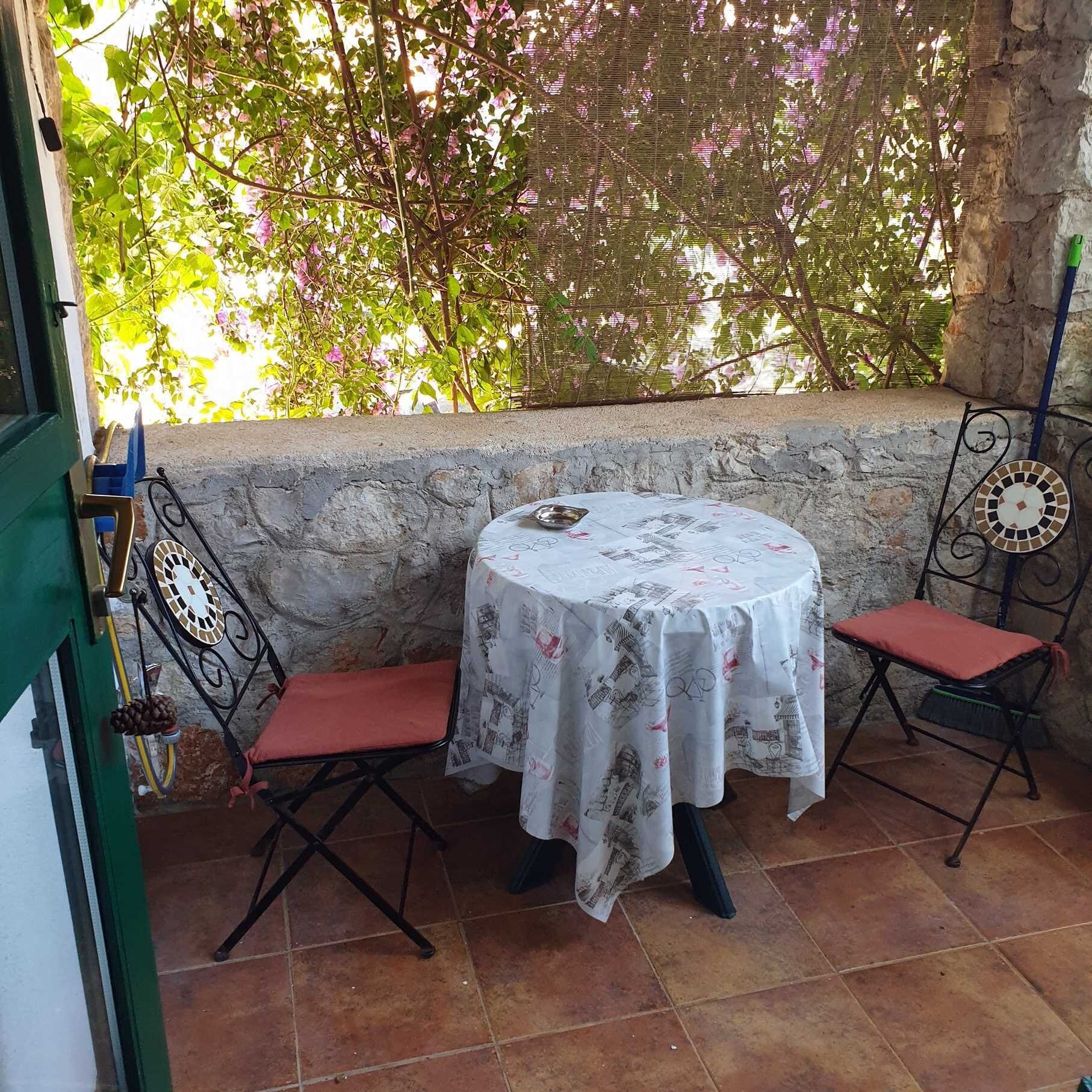 Um pequeno pátio ao ar livre com uma mesa redonda e duas cadeiras, uma parede de pedra e vista para o verde.