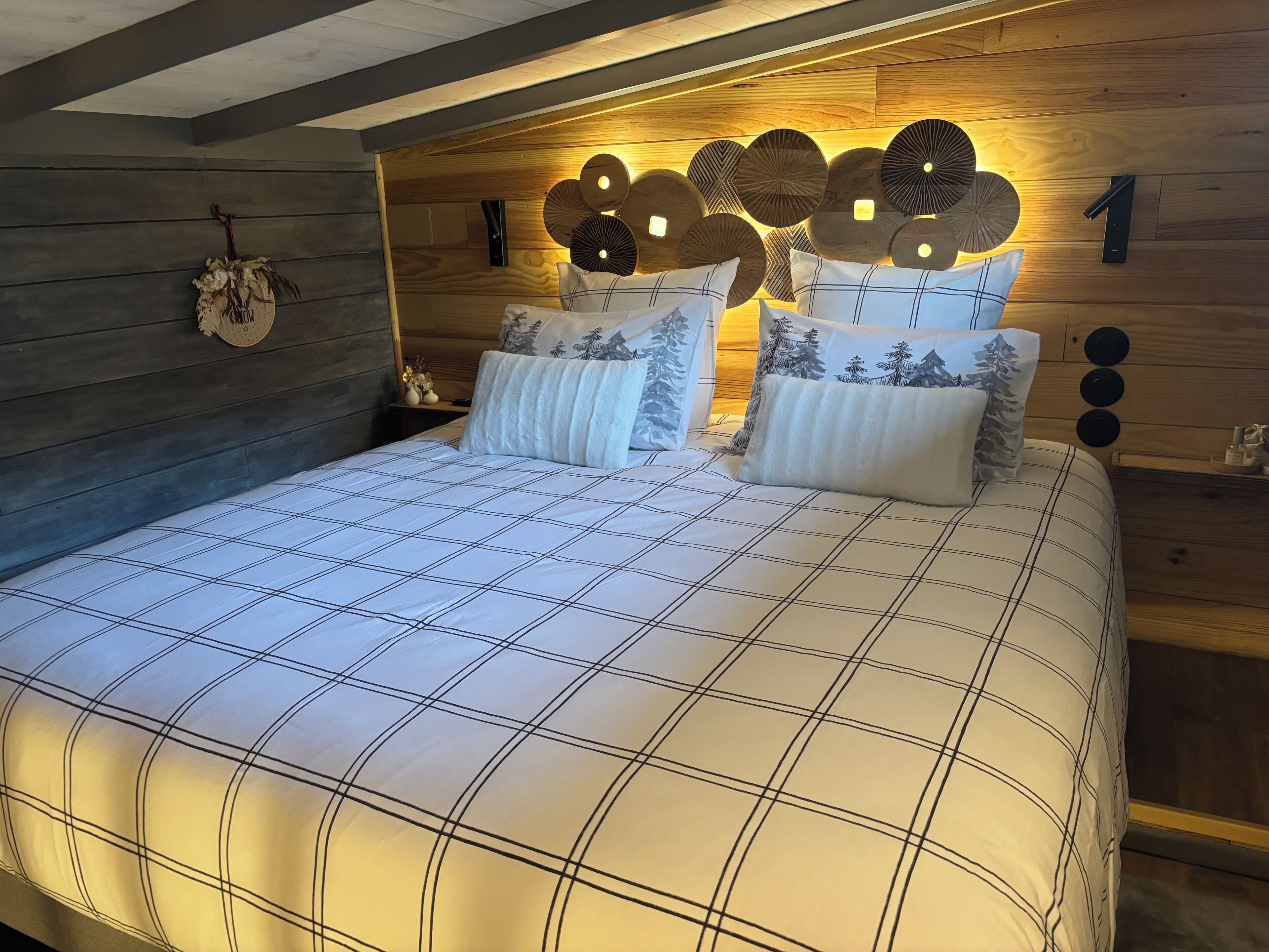 Una cama bien hecha con una colcha a cuadros y varios almohadones, un cabecero de madera con adornos y una mesita de noche con una lámpara pequeña y un objeto decorativo.