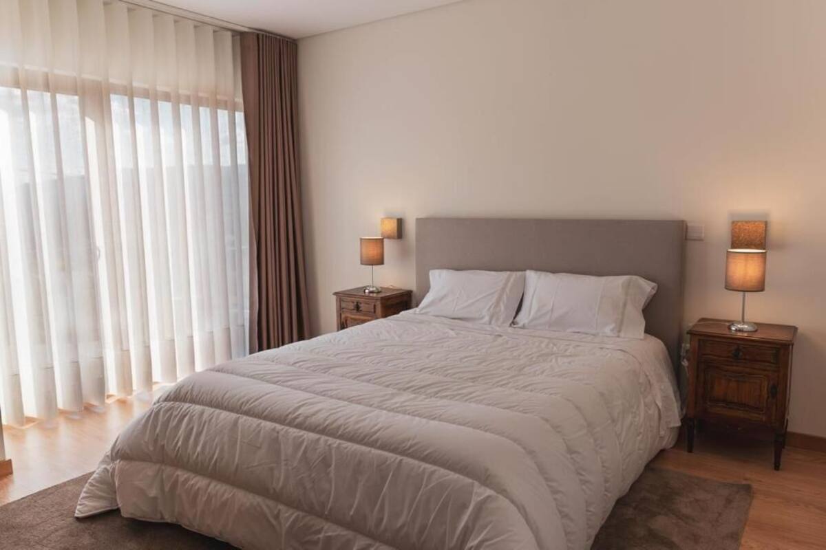 Ein ordentlich bezogenes Bett mit beiger Bettdecke und Kissen, das von zwei Nachttischen mit Lampen eingerahmt wird, in einem Zimmer mit durchsichtigen Vorhängen und einem großen Fenster.