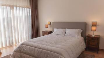 Una cama bien hecha con edredón color beige y almohadas, flanqueada por dos mesitas de noche con lámparas, en una habitación con cortinas translúcidas y un ventanal grande.