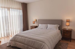 Una cama bien hecha con edredón color beige y almohadas, flanqueada por dos mesitas de noche con lámparas, en una habitación con cortinas translúcidas y un ventanal grande.
