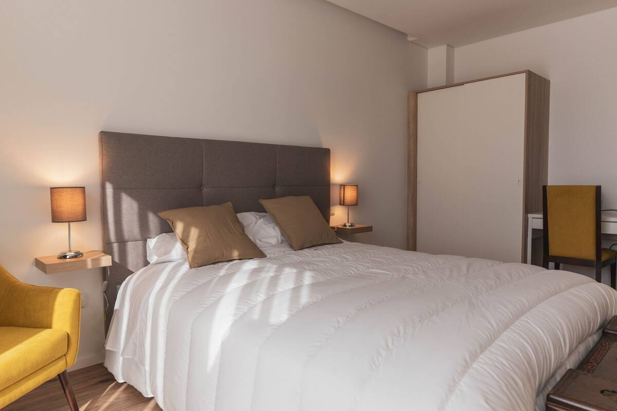Ein modernes Hotelzimmer mit einem großen Bett, zwei Nachttischlampen, einem Sessel und einem Kleiderschrank.