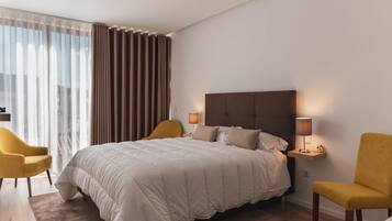 A modern hotel room with a large bed, two bedside lamps, a nightstand, a chair, and a view of the city through sheer curtains.