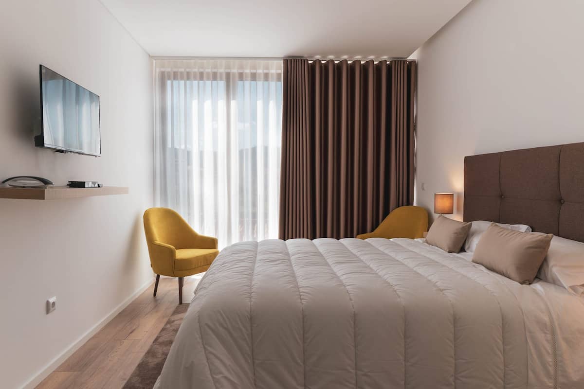 Ein modernes Hotelzimmer mit einem großen Bett, einem flachbildfernseher an der Wand, einem kleinen Schreibtisch mit Lampe und einem Fenster mit durchscheinenden Vorhängen und einem braunen Vorhang.