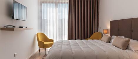 A modern hotel room with a large bed, a flat-screen TV mounted on the wall, a small desk with a lamp, and a window with sheer curtains and a brown drape.