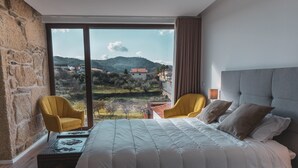 A bedroom with a large bed, two yellow armchairs, a view of a mountainous landscape, and a stone wall.
