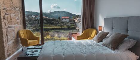 A bedroom with a large bed, two yellow armchairs, a view of a mountainous landscape, and a stone wall.