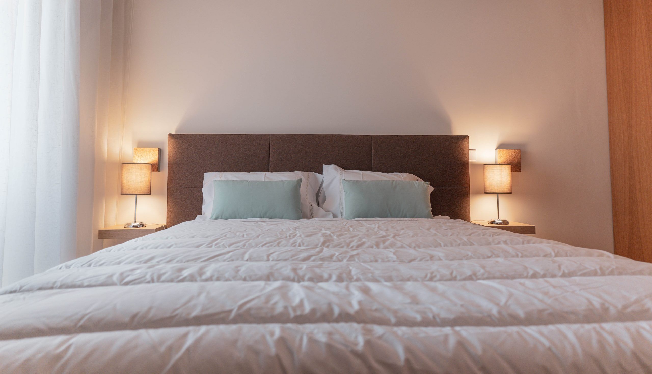 A neatly made bed with a brown headboard, two bedside lamps, and a window with curtains.