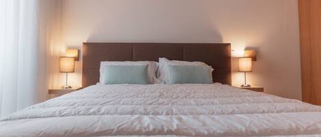 A neatly made bed with a brown headboard, two bedside lamps, and a window with curtains.