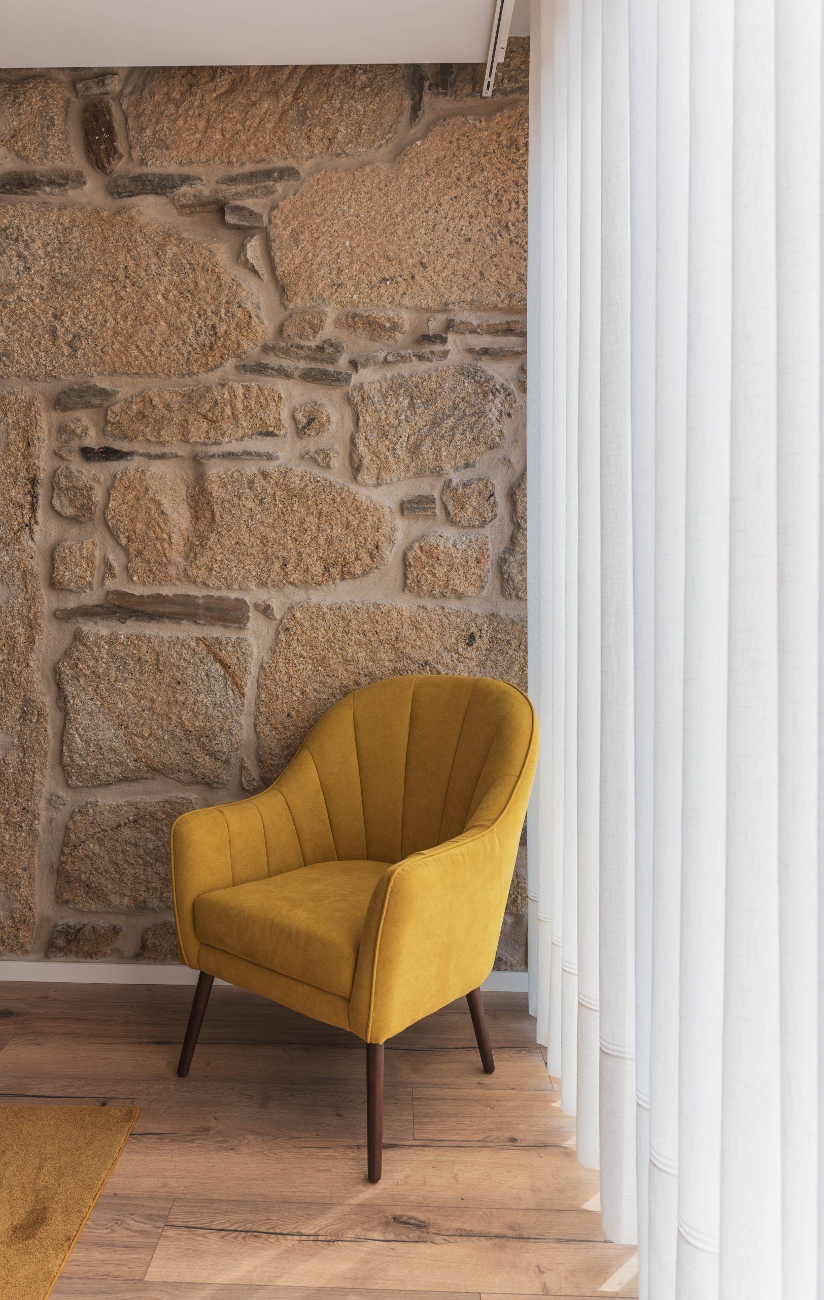 Un fauteuil de couleur moutarde placé devant un mur de pierre, avec un plancher en bois et des rideaux blancs.