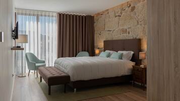 Una habitación de hotel moderna con una cama grande, una pared decorativa de piedra, vistas a través de ventanas del suelo al techo y una zona de estar con una silla y una lámpara.