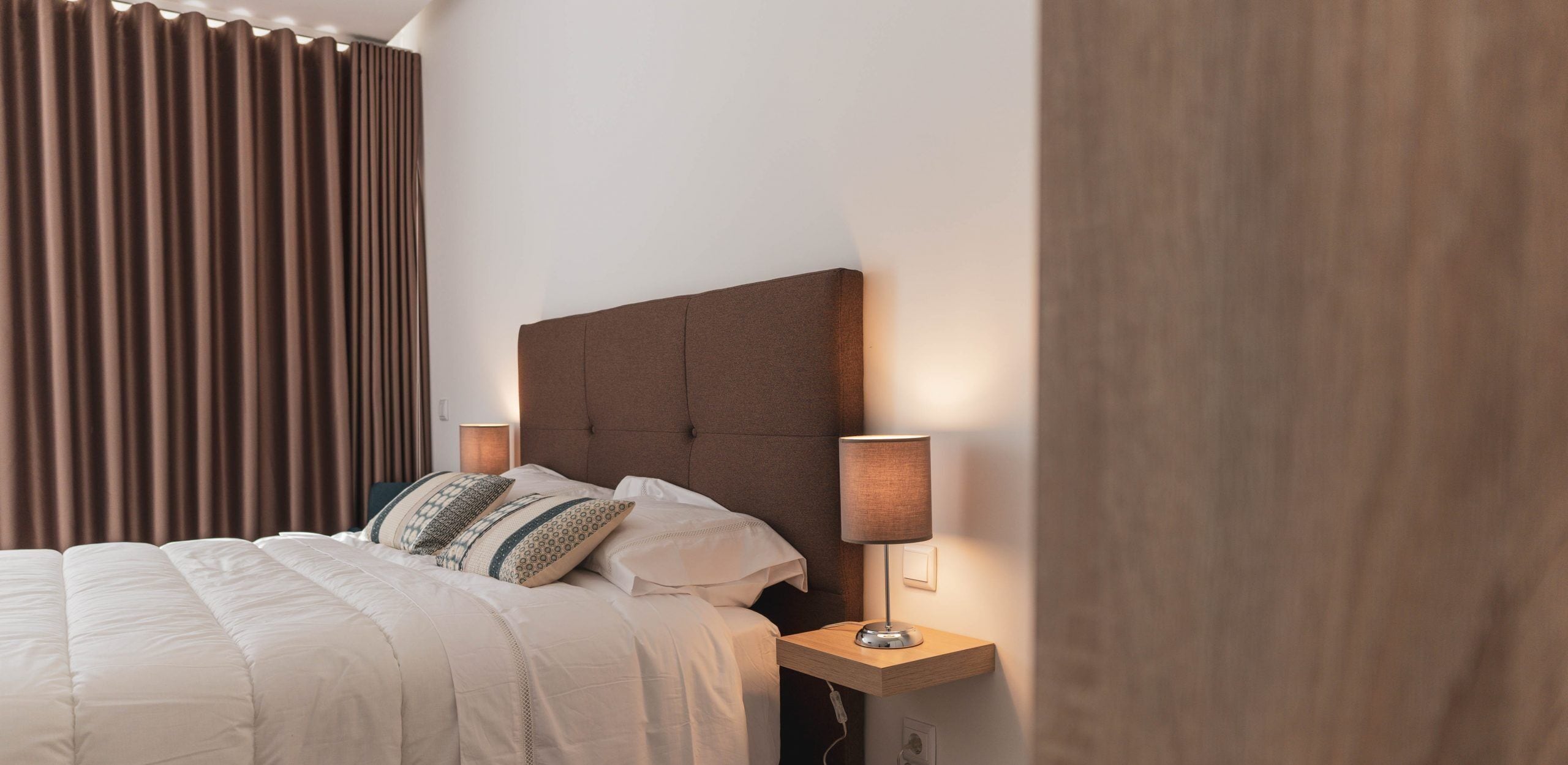A neatly made bed with a headboard, two bedside lamps, and a window with curtains.