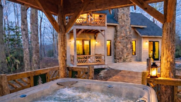 Un bain à remous situé sur une terrasse en bois, avec un pilier en pierre, une balustrade en bois et une maison en arrière-plan.