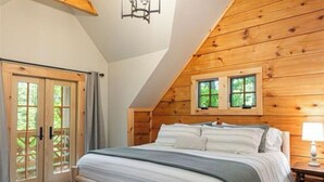 Une chambre avec un plafond en pente, des poutres en bois, un lit avec un cadre en bois, une table de chevet et une fenêtre donnant sur un paysage verdoyant.