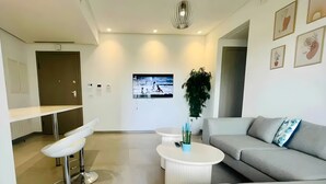 Un salon moderne avec un canapé gris, une table basse blanche et un tabouret de bar blanc. Un téléviseur est fixé au mur, il y a une plante en pot et une porte qui donne sur une autre pièce.