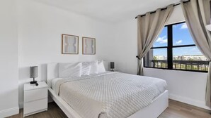 A bedroom with a large bed, a nightstand, two framed pictures on the wall, and a window with curtains. - INTERNATIONAL dr/UNIVERSAL 2 BEDROOMS KING SUITE with free parking  (Orlando)