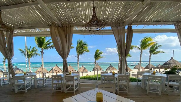 Een terras buiten met tafels en stoelen, met uitzicht op een strand met palmbomen en rieten parasols.