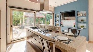Een moderne keuken met een groot keukenblok, een gasfornuis en een spoelbak. Er zijn twee barstoelen en een eettafel gedekt voor twee personen. De keuken heeft een blauwe accentmuur en een groot raam met uitzicht op een tuin.
