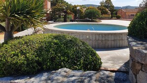Une piscine entourée de murs de pierre et d’une végétation luxuriante, avec un ciel bleu clair et des collines lointaines en arrière-plan.