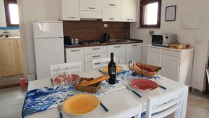 Une salle à manger avec une table blanche dressée pour quatre personnes, entourée de chaises blanches. On aperçoit au fond une cuisine avec des meubles blancs et un four à micro-ondes posé sur le plan de travail.