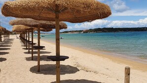 Une plage bordée de rangées de parasols en chaume et de tables en bois, une mer d’un bleu limpide et une côte lointaine parsemée de bâtiments.